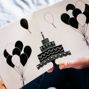 open baby board book showing a black and white picture of a cake and balloons